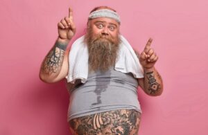 Vertical shot of bearded Caucasian adult man gets sweaty during his training session, works hard on losing excess weight and points above. Funny obese guy wears headband shirt, goes in for sport