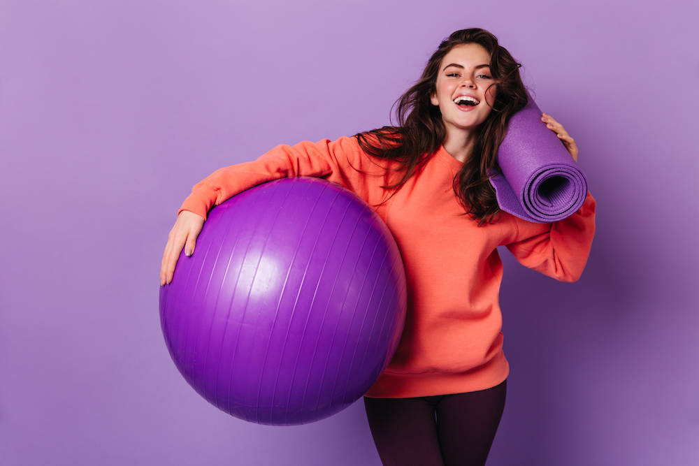 Beautiful woman in leggings and bright sweatshirt is smiling and posing with purple mat and fitball.