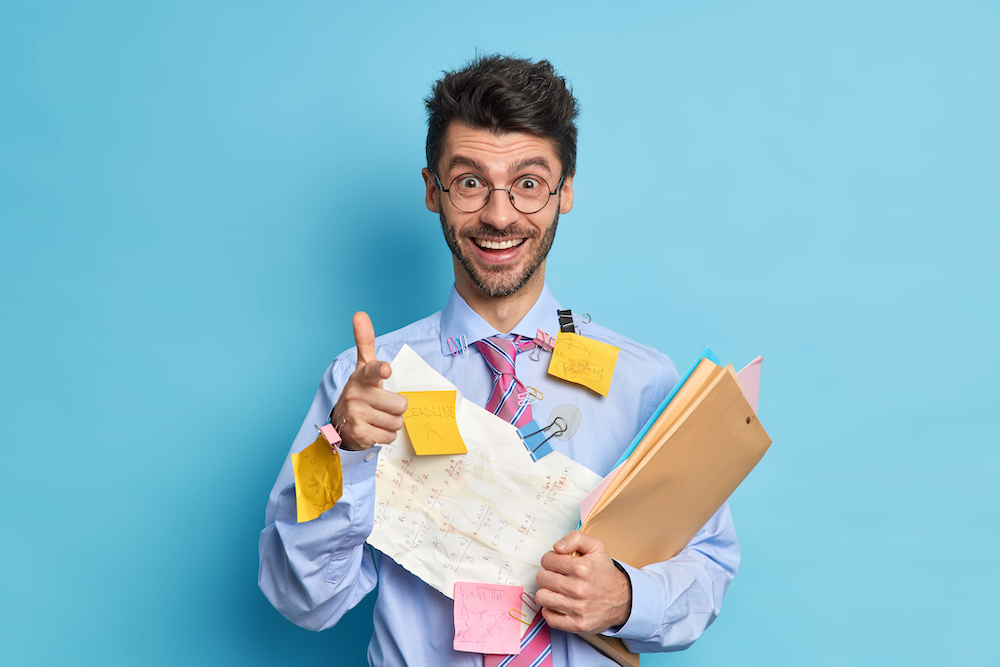 Cheerful young man coworker happy to finish project work covered with papers and stickers points at you makes finger gun gesture. Successful dilligent student busy doing course work poses indoor