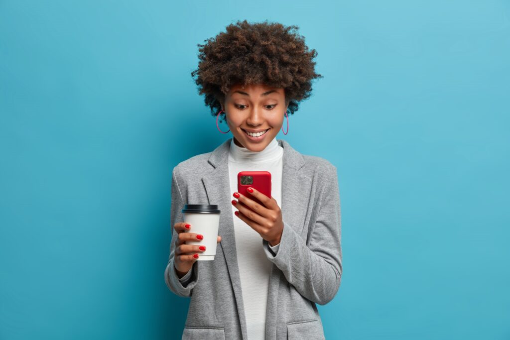 Positive Afro American woman in formal wear focused in smartphone device, reads online news and drinks take away coffee. Successful businesswoman enjoys communicating with friends in social networks