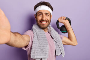 Bodybuilding concept. Glad unshaven man does sport with weight, wears headband, casual t shirt uses towel makes selfie portrait for sharing photos in social networks. Male has training with kettlebell