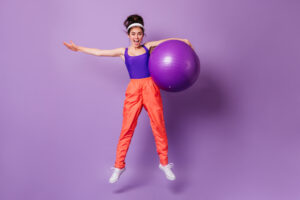 Cheerful young girl in purple sports top and bright pants jumps with fitball on isolated background