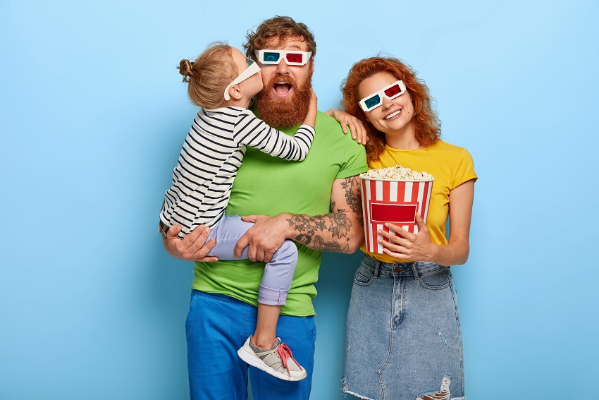 Ginger family choose pleasant way of spending free time, book tickets in advance for premier of movie, small daughter kisses and embraces father, mother holds bucket with popcorn, being happy