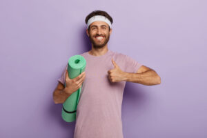 Glad unshaven man makes okay gesture, keeps thumb up, holds rolled up karemat, shows he liked yoga training wears casual t shirt in one tone with background. People, workout, healthy lifestyle concept