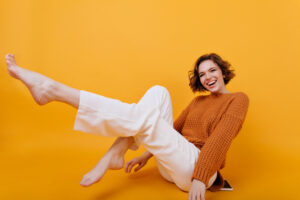 Smiling barefooted girl with eacstatic face expression posing with legs up. Laughing female model in woolen sweater chilling during indoor photoshoot..